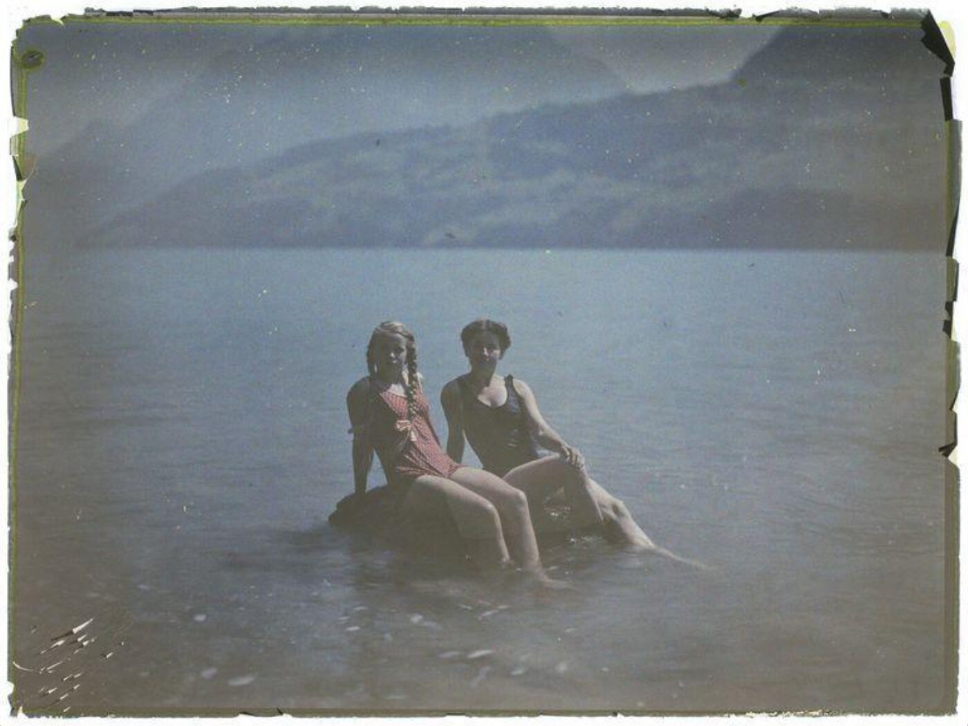 Two young women seated on a rock in the sea. The colours of the image have changed over time.