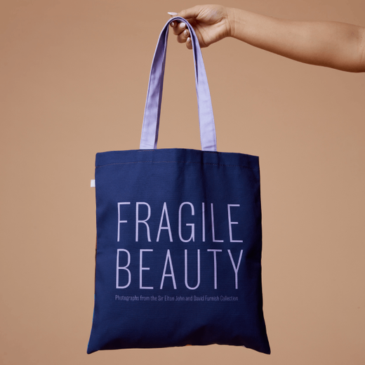 A blue tote bag with a purple text spelling 'Fragile Beauty' is held up from its handle.