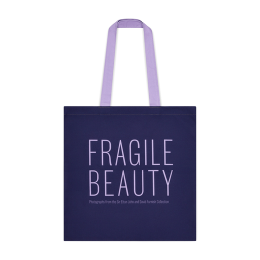A blue tote bag with a purple text spelling 'Fragile Beauty' in capital letters and featuing a lilac handle.
