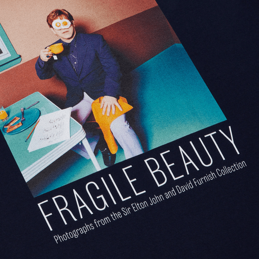 A detail of a blue t-shirt featuring Elton John sitting at a dining table with fried eggs behind his glasses and the text 'Fragile Beauty' in capital letters.