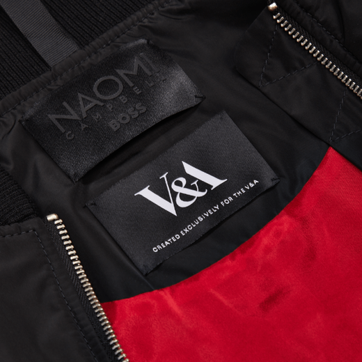 A detail shot of the collar of a black jacket with red inner lining. Two black labels show the name Noami Campbell and the V&A logo, respectively.