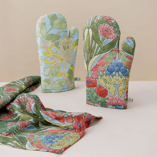 Two oven gloves with floral patterns are propped up on a table against a cream background.
