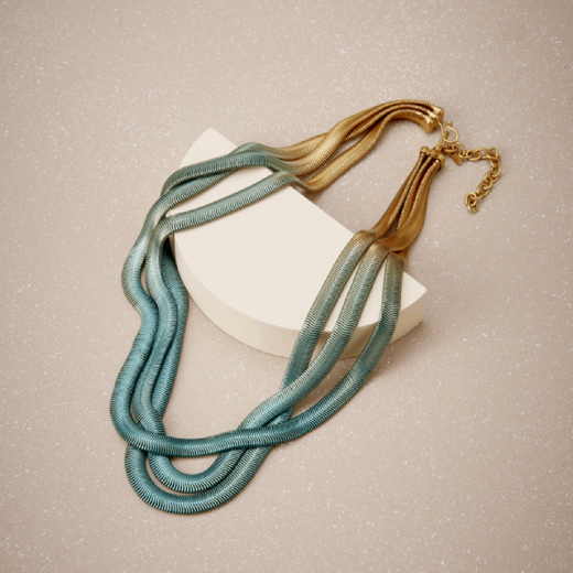 A chain necklace in a gold and blue colour gradient. The necklace is placed on a cream plinth and light brown background.