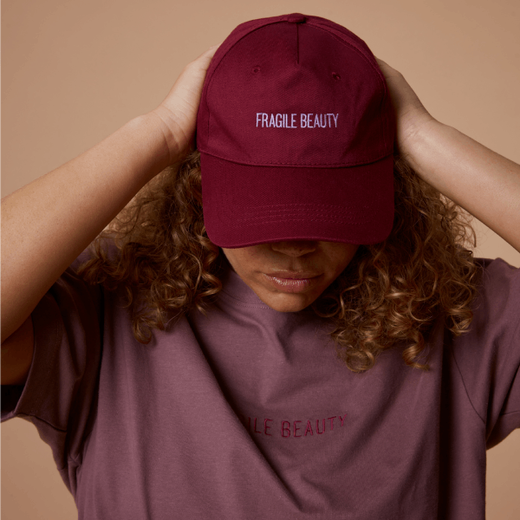 A woman wearing a burgundy cap with embroidered text spelling Fragile Beauty on the front.