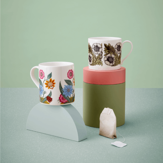 Two china mugs with floral patterns are displayed on small plinths against a green background.