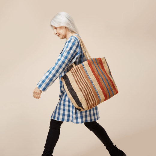 A woman wearing a checked blue shirt and black trousers walks carrying a large striped handbag.