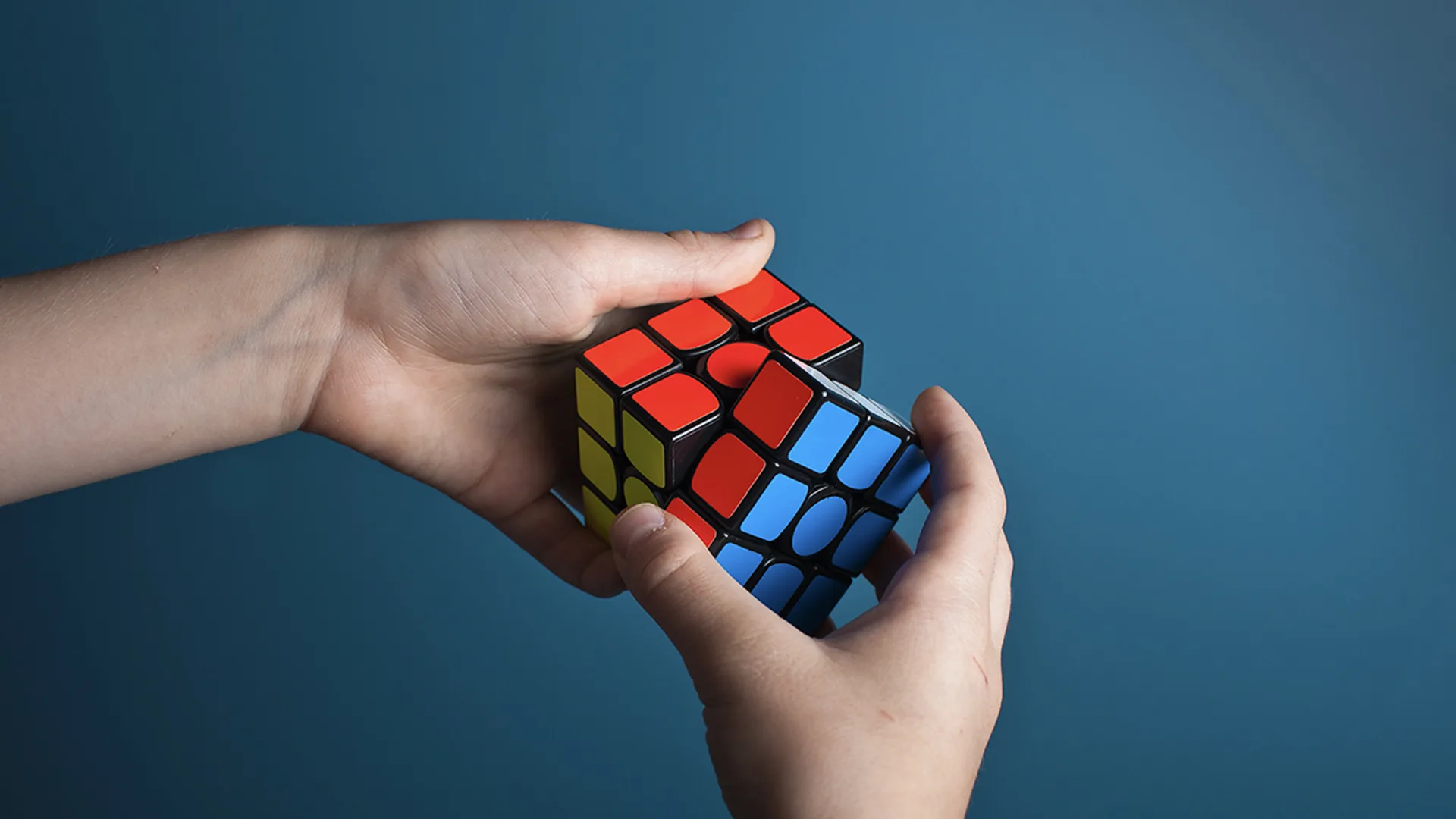 Close-up of hands holding and trying to solve Rubik's cube on plain turquoise background.