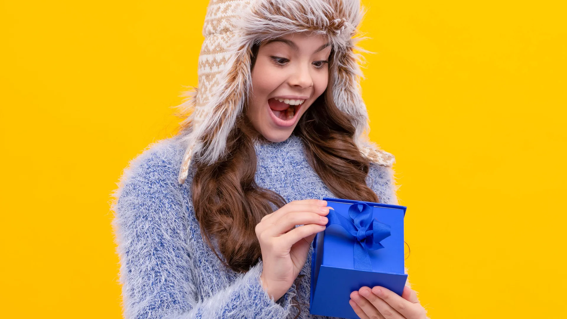 Photograph of a girl wearing a furry hat and jumper opening a present on a yellow background