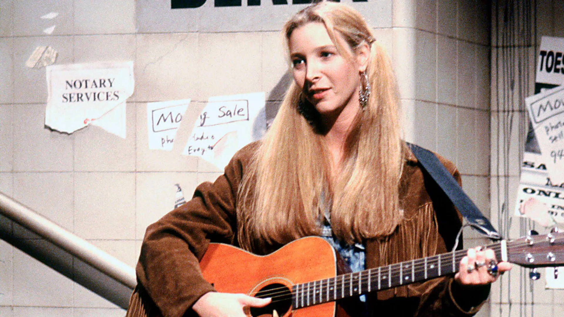 A photograph of the character Phoebe in Friends with a guitar against a stone wall with posters on it