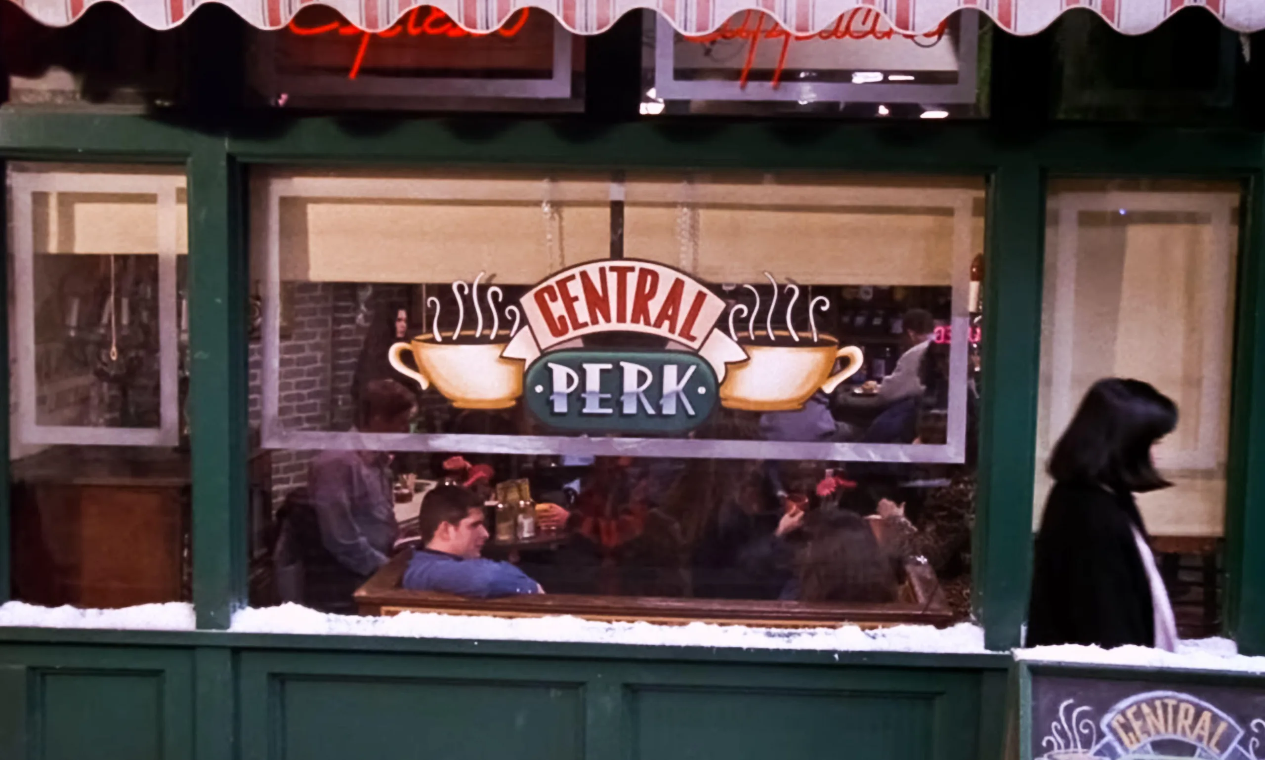 A photograph of the coffee shop in Friends that has a sign "central park" with green frames and snow on windowsill. People are inside sitting on the sofas.