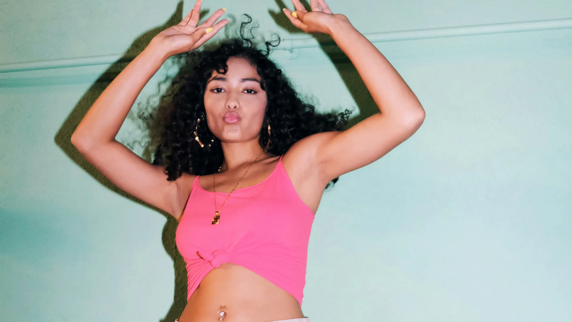 A photograph of a woman with dark hair wearing a pink top looking directly at the camera with her arms raised as she is dancing against a blue wall background