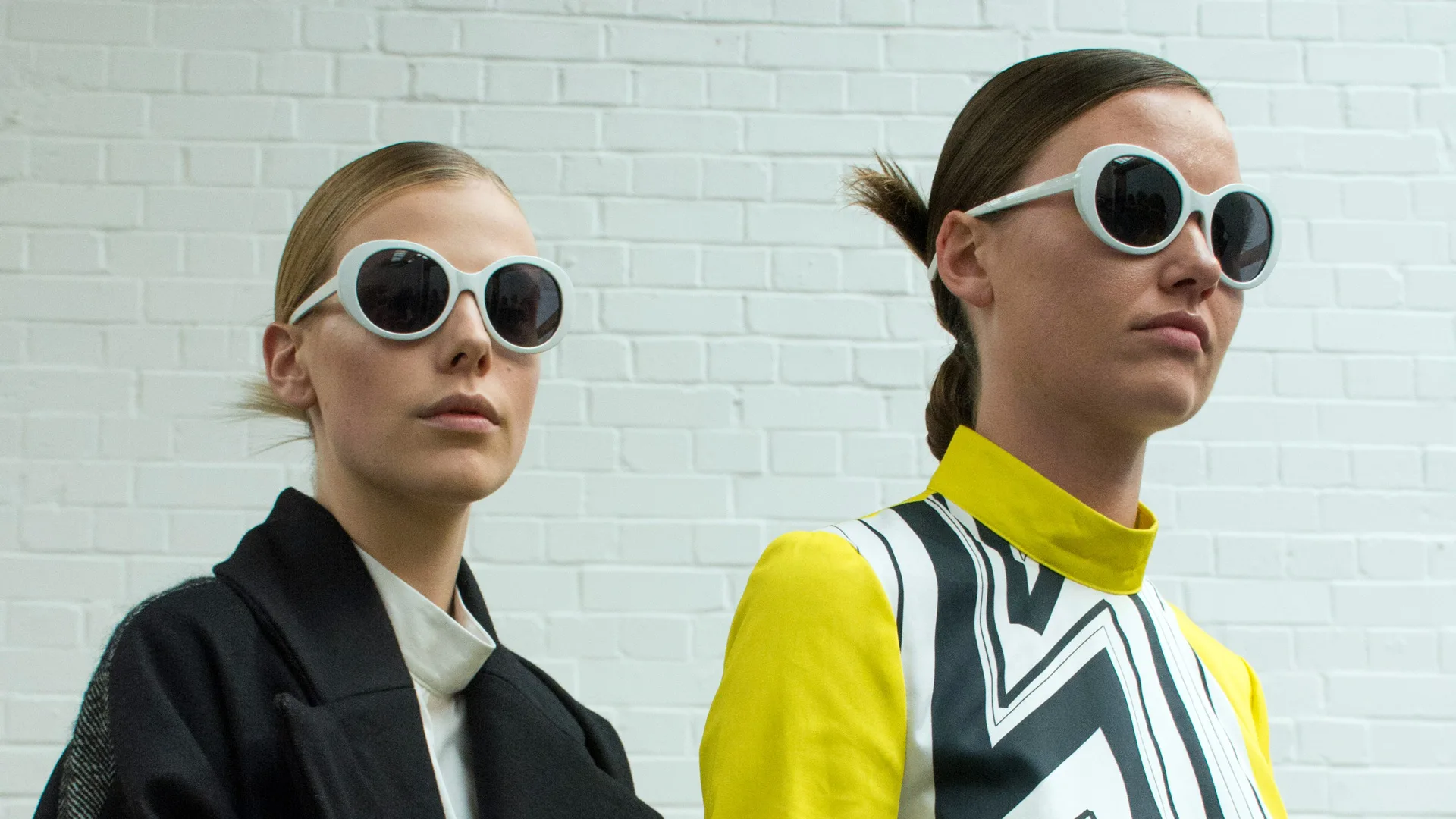 Two women with sunglasses stood against a white painted brick wall