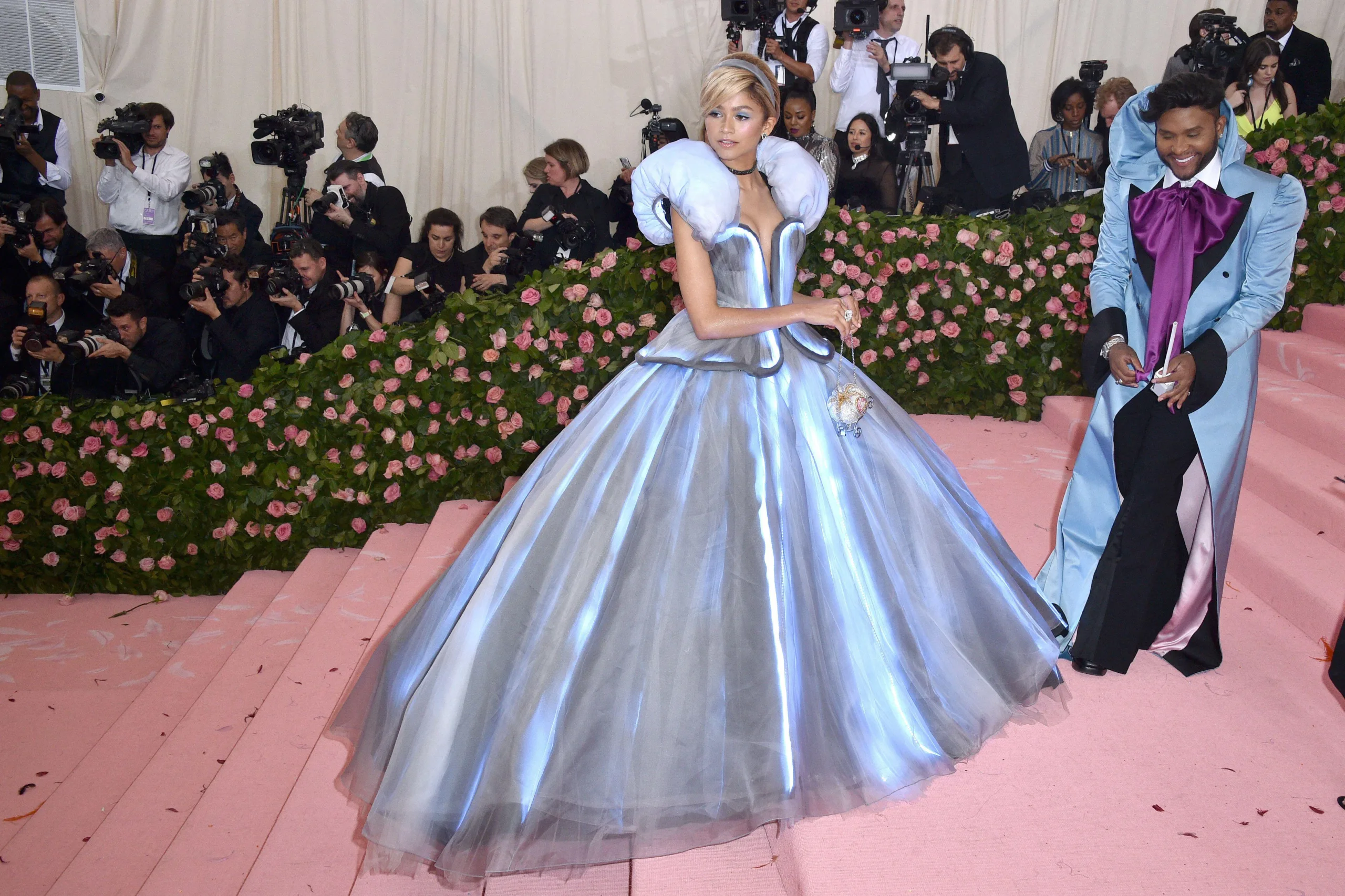 A photo of Zendaya at the Met Gala 2019 wearing a light up Cinderella dress as she walks up the stairs with people beside her