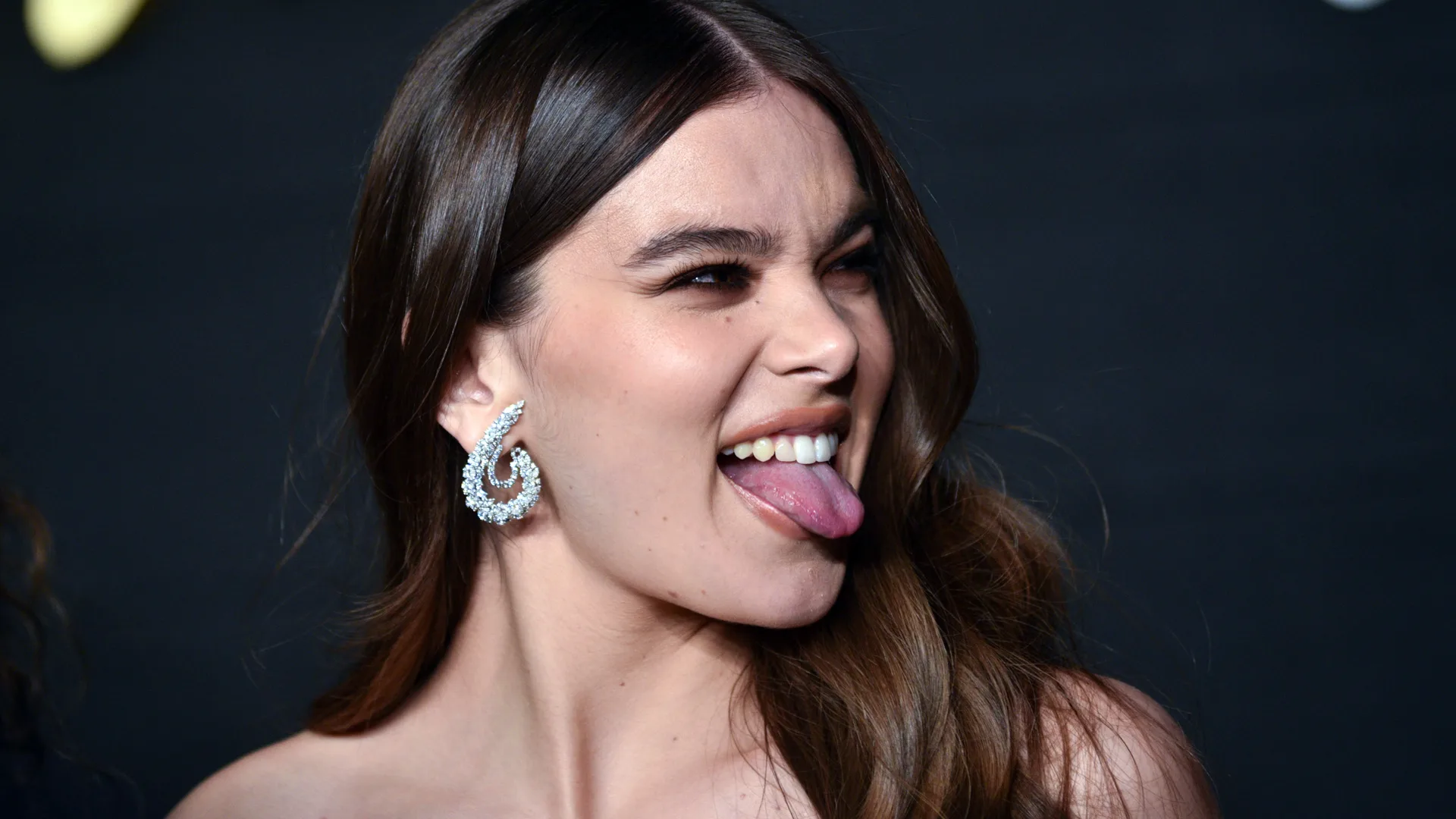 A photograph of the singer Hailee Steinfeld sticking her tongue out to someone off camera in a cheeky smile. She is wearing silver curly earrings with her brown hair down over her shoulder against a black background.