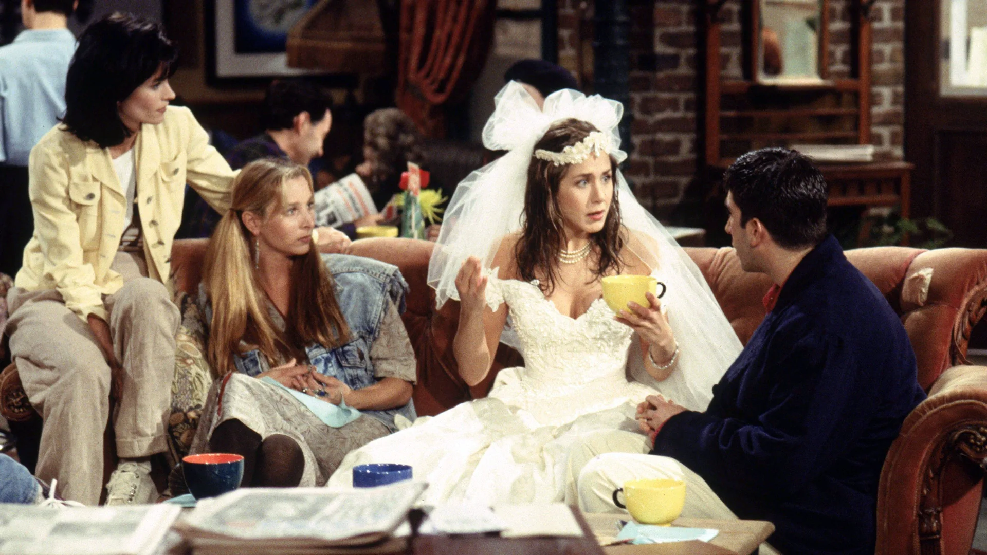 A photograph of a scene from the TV show Friends showing Monica, Phoebe, Rachel and Ross sat in the coffee shop looking concerned. Rachel is wearing a wedding dress and holding a yellow cup. The background has a brick wall.