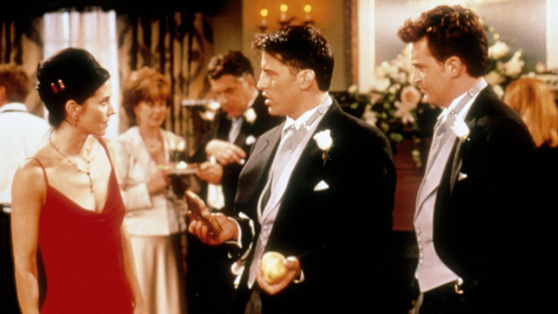 A photograph of Monica, Joey and Chandler in a scene from the TV show Friends all wearing evening wear having what looks like a tense discussion. Monika is in a red dress and Joey and Chandler are in dinner suits. Behind them are two people looking at an invite.