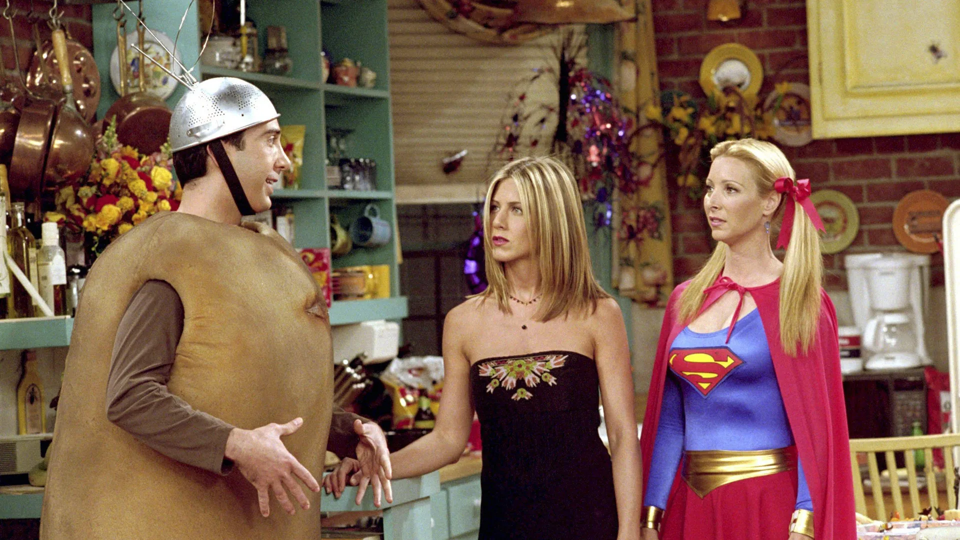 A photograph of the cast of Friends - Ross, Rachel and Phoebe in Halloween costume. Ross is dressed like a potato with a silver colander on his head, Rachel is in a black dress and Phoebe is dressed as Superwoman. They are stood in a kitchen.