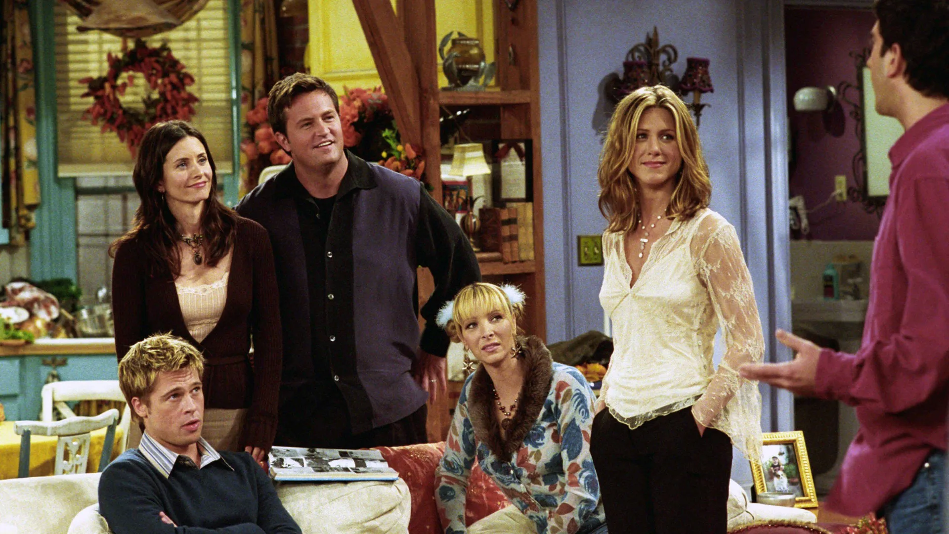 A photo of a scene from the TV show Friends where all of the characters are stood listening to something Ross is saying in their apartment