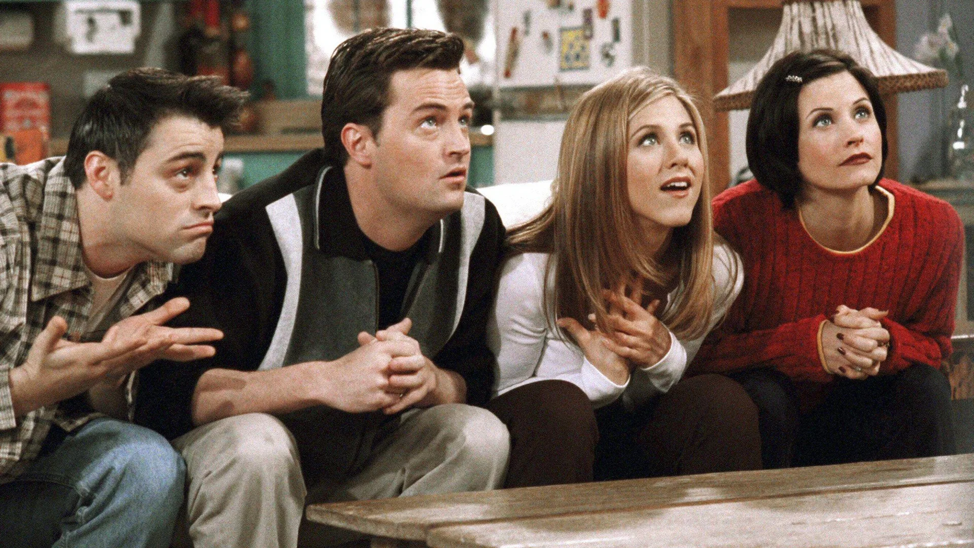 A photograph of the Friends characters Joey, Chandler, Rachel and Monika sat on a sofa looking up at something or someone half surprised.