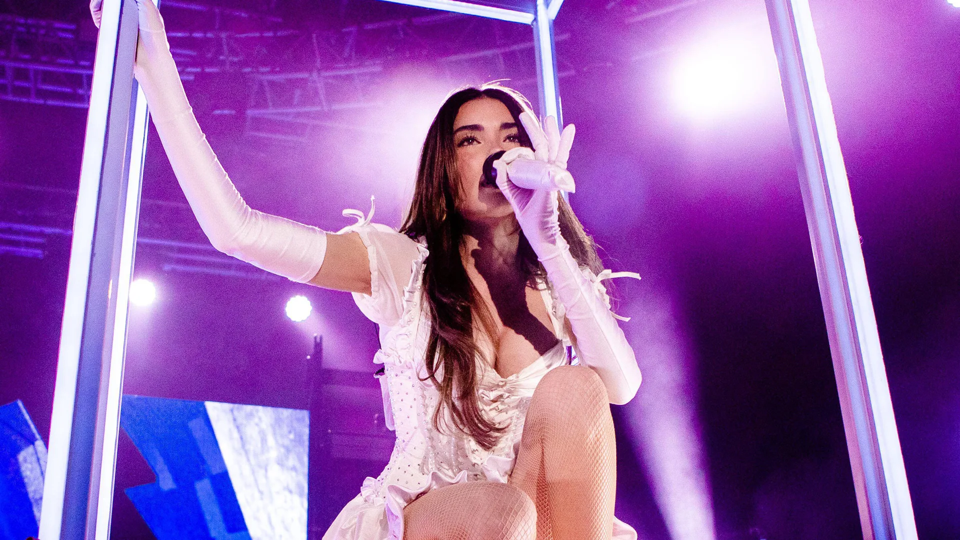 A photograph of the singer Madison Beer performing on stage inside a light box structure. She is holding a microphone, wearing white gloves and a white dress with her hair long and brown. The background is stage lights with purples and blues.
