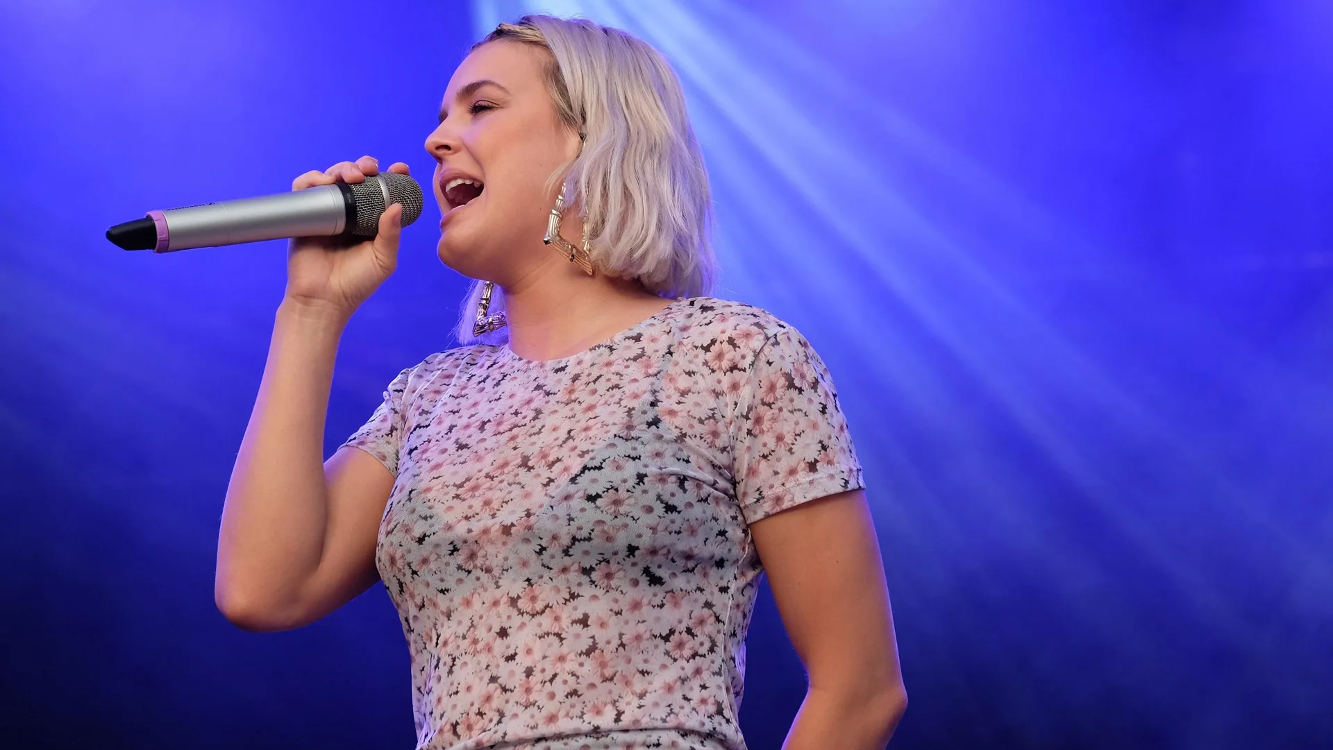 A photograph of the singer Anne-Marie Nicholson performing at Bestival. She is wearing a floral mesh top, singing into a microphone with blonde bobbed hair, against a purple background with a stage light coming down on her.