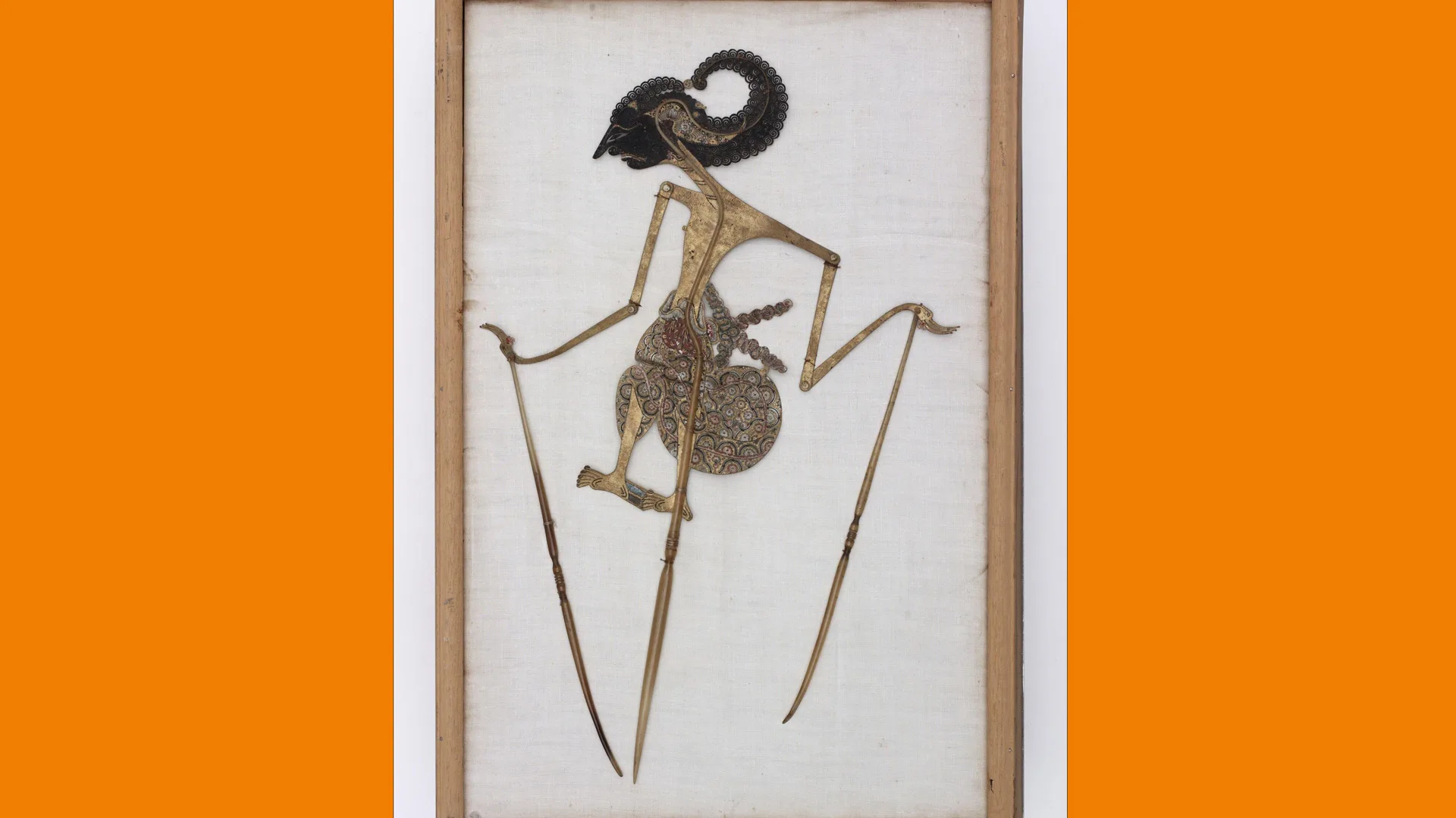 A photo of a very delicate shadow puppet with long legs and arms against a grey background with orange borders