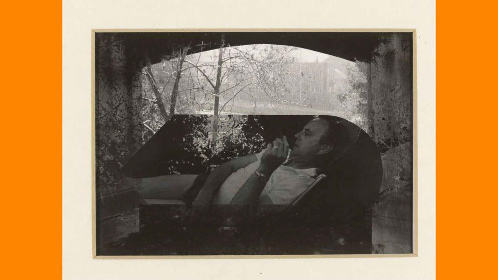 A black and white moody photograph of a man lying on a sofa in a white top against a window with trees in the background