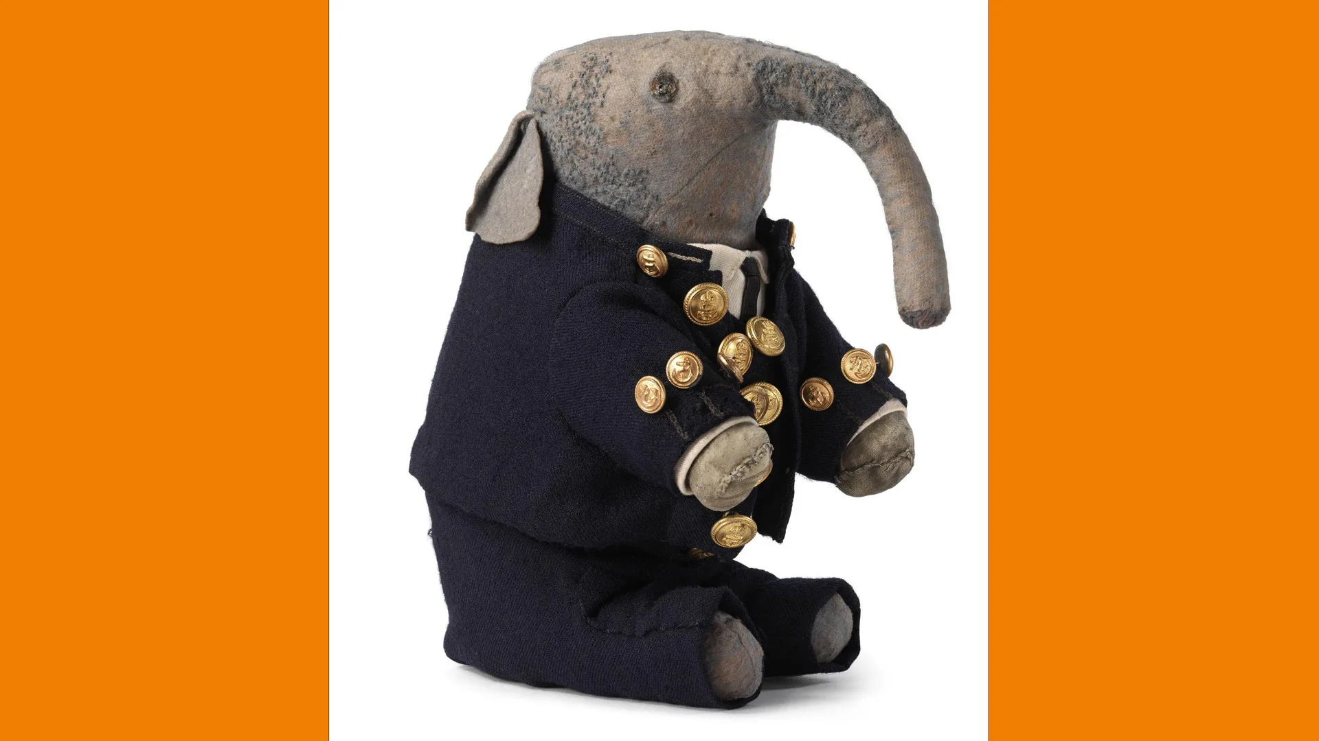 A photograph of a Victorian child's soft toy elephant called Pumpie. Pumpie wears a sailor suit in navy with gold buttons and has grey fur. He is in a seated position against a white background with orange borders.