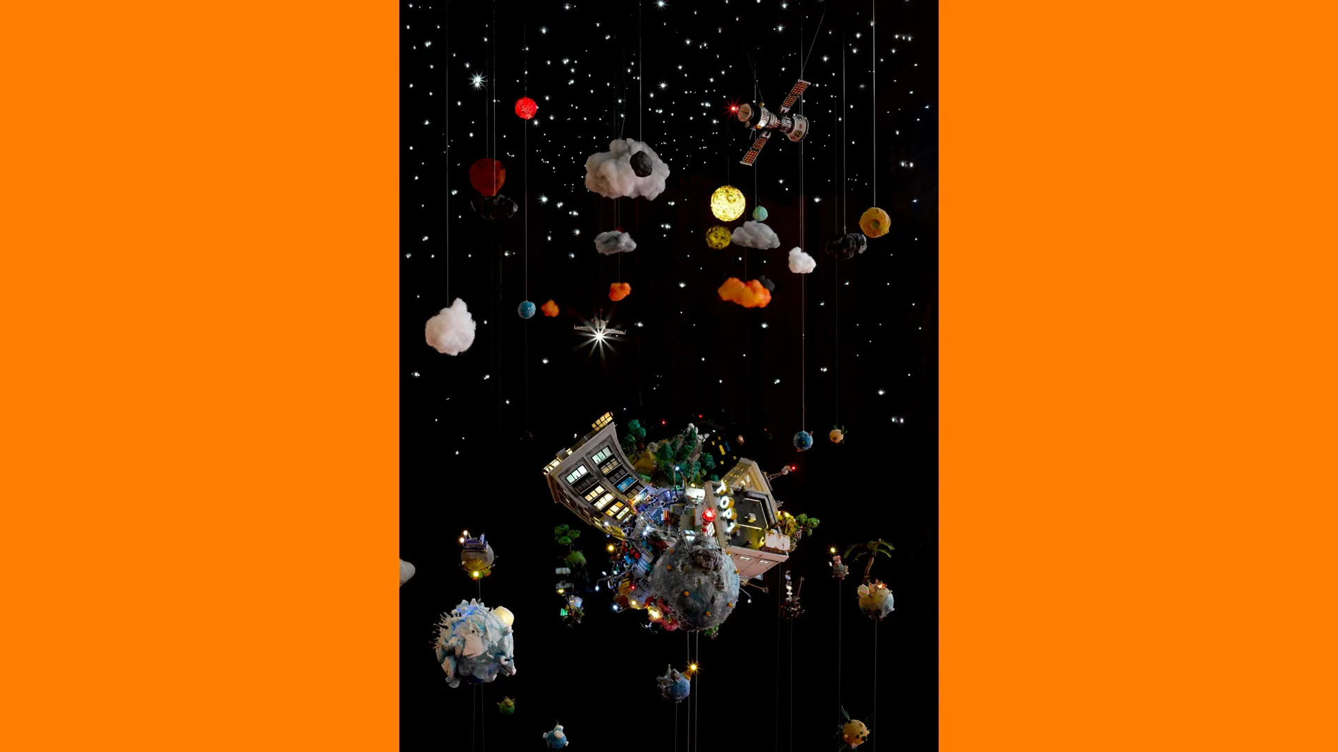 A photo of a delicate art installation depicting planet earth and the ecology of earth set in space. There are 30 pieces made of different materials all hanging from string. The structure is set against a black background with orange borders,