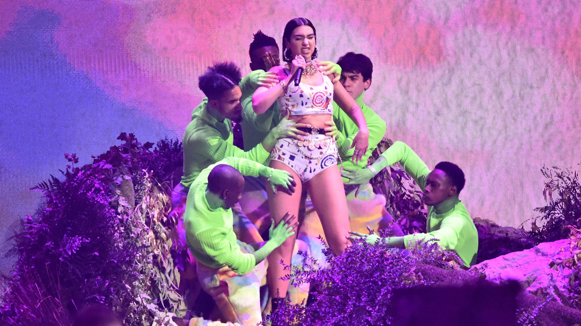 A photo of Dua Lipa performing at the 2019 BRIT awards wearing a cropped hotpants and top combo with dancers surrounding her in neon green outfits against a pink and purple stage background.