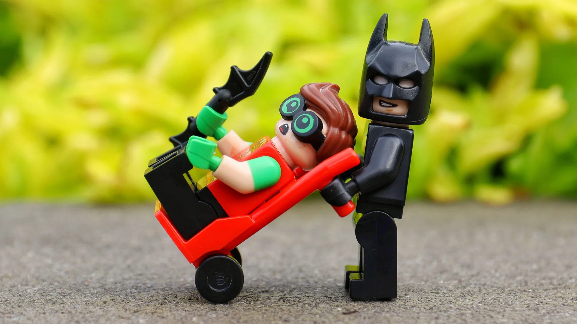 A photograph of two LEGO figures - Batman and Robin. Batman is holding Robin in a trolley and Robin is holding a bat. They are stood on gravel with green leaves in the background.