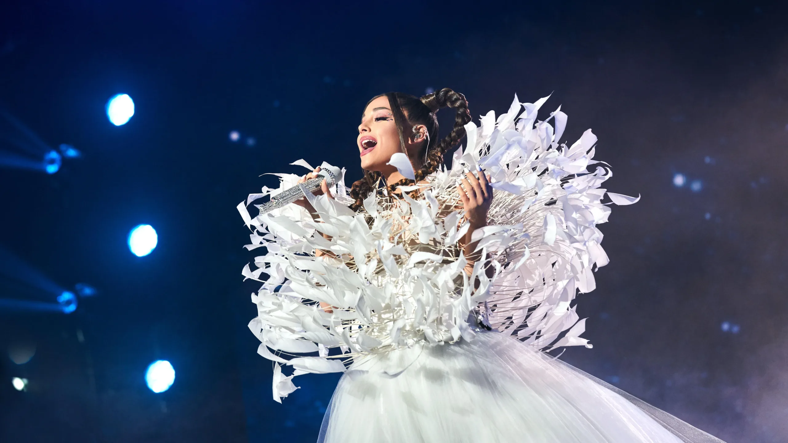 Ariana Grande sings on stage wearing white feathered outfit