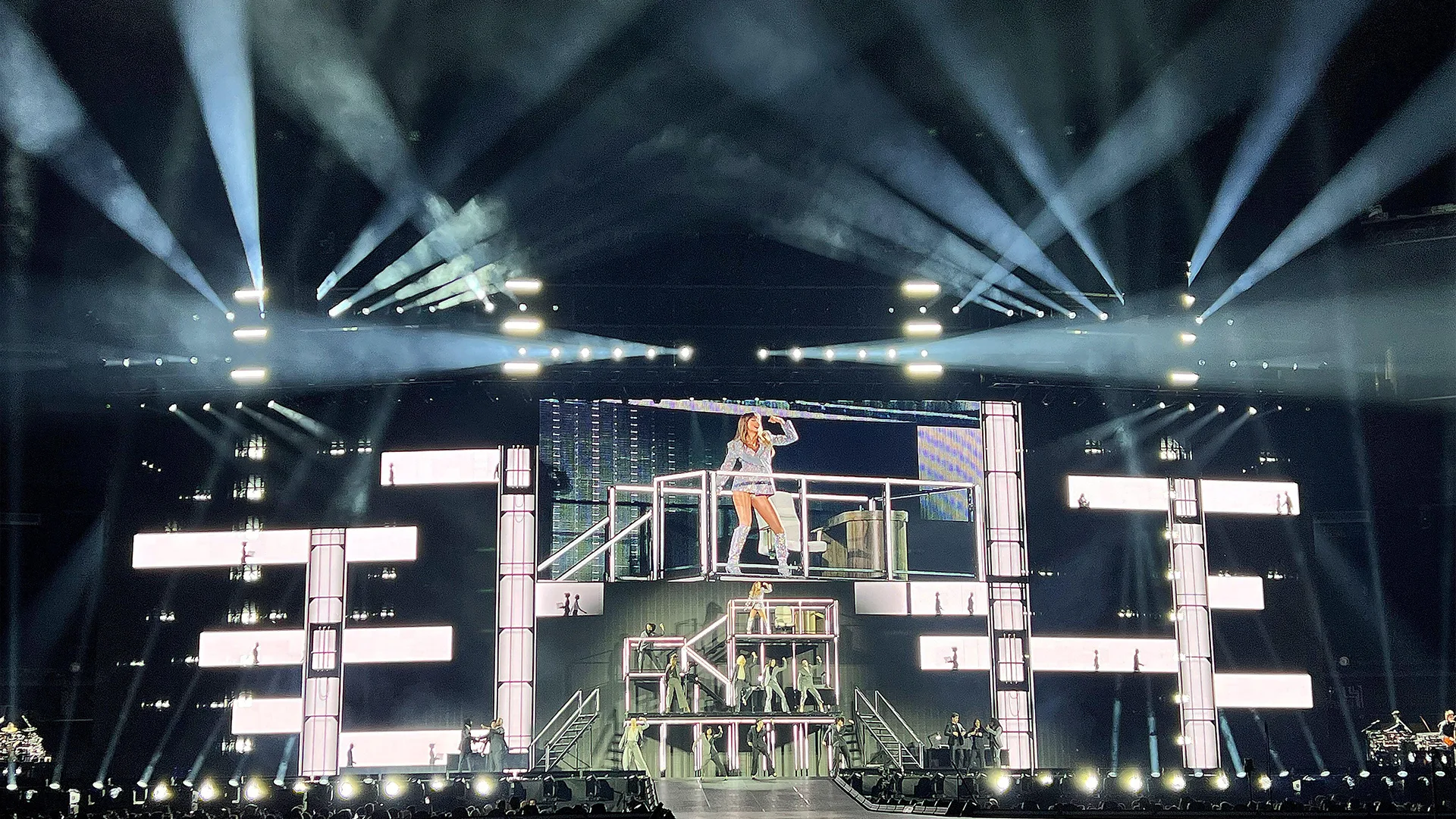 Taylor Swift singing on The Eras Tour stage. The stage design shows is a black background with blocks of white windows and silhouettes of people in them