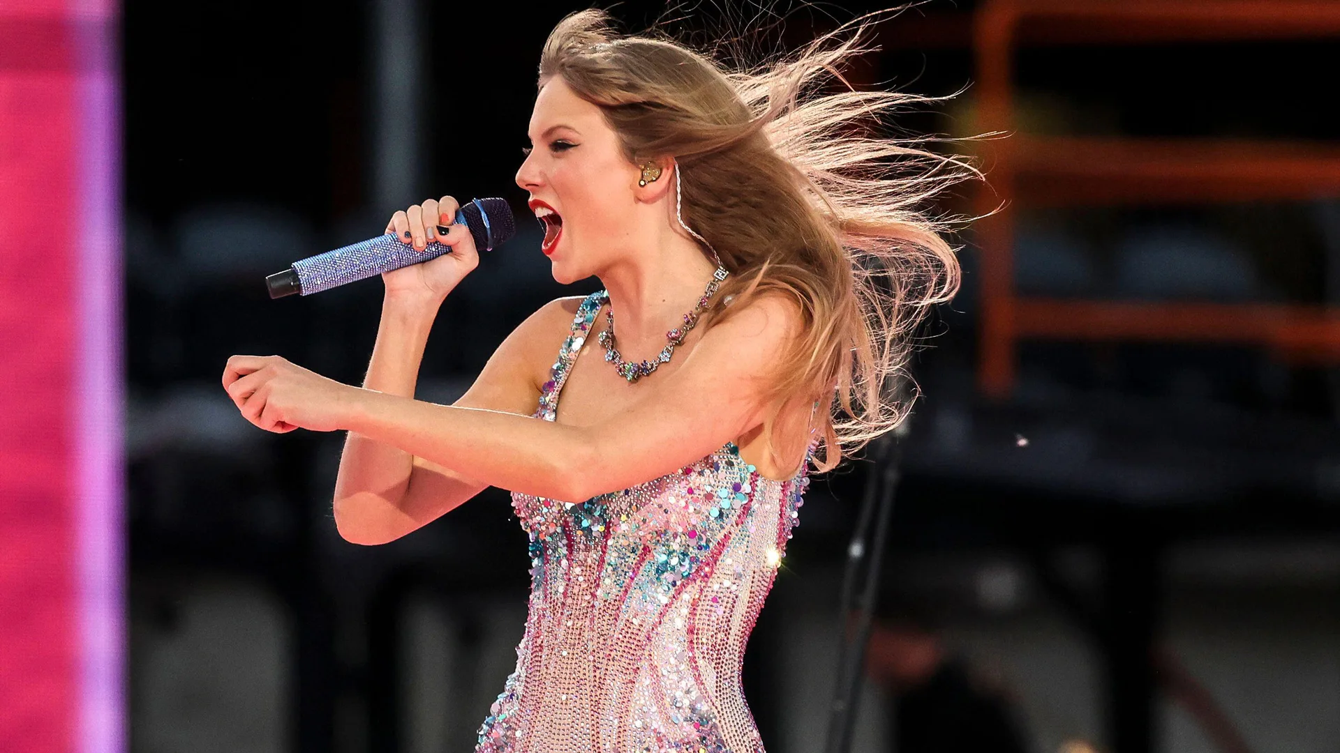 A photograph of Taylor Swift singing into the mic at her Eras Tour. She is wearing a crystal bodysuit and her hair is being blown.