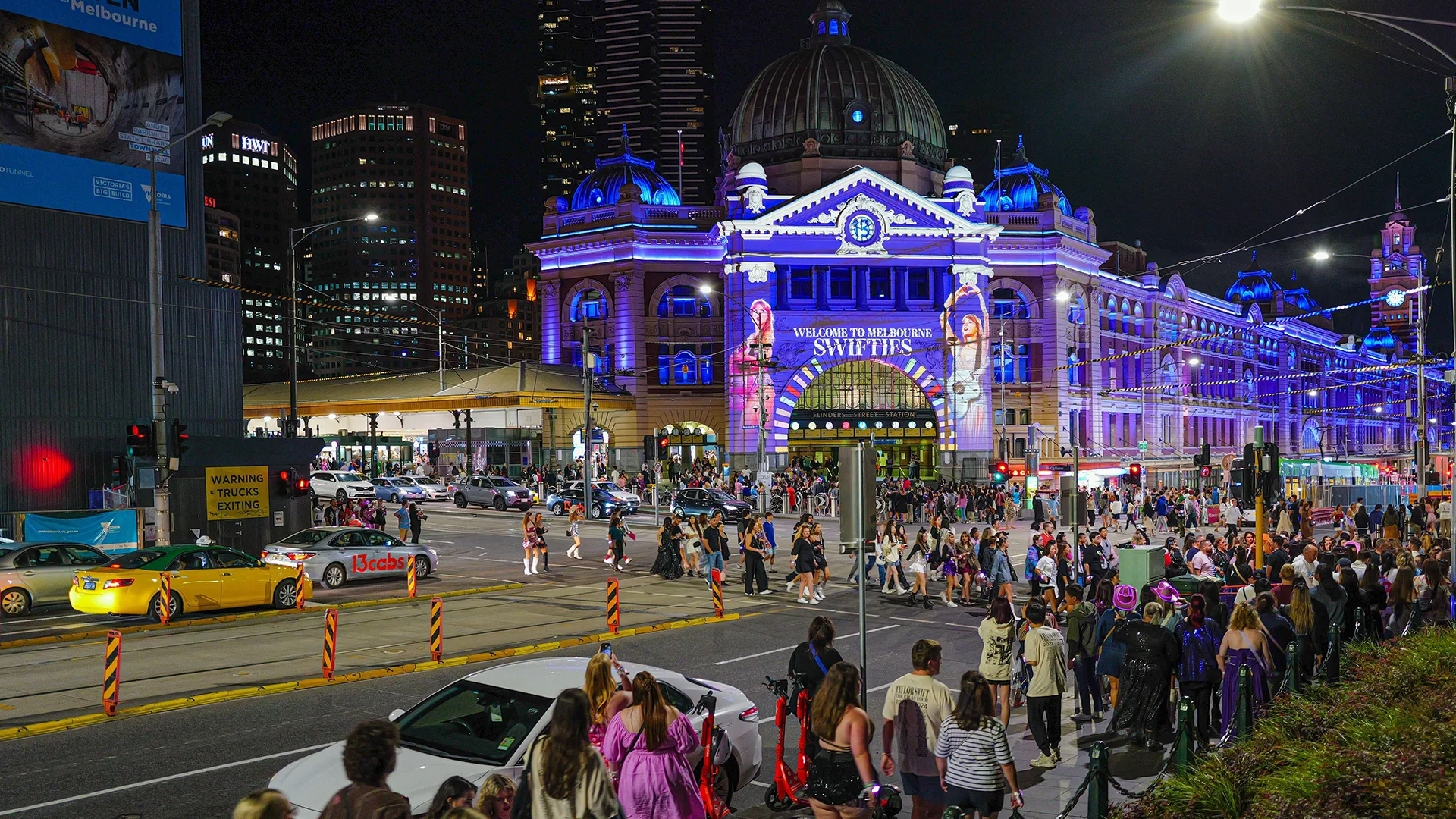Fans queue outside large concert venue in Melbourne that has the Eras concert poster projected onto the building