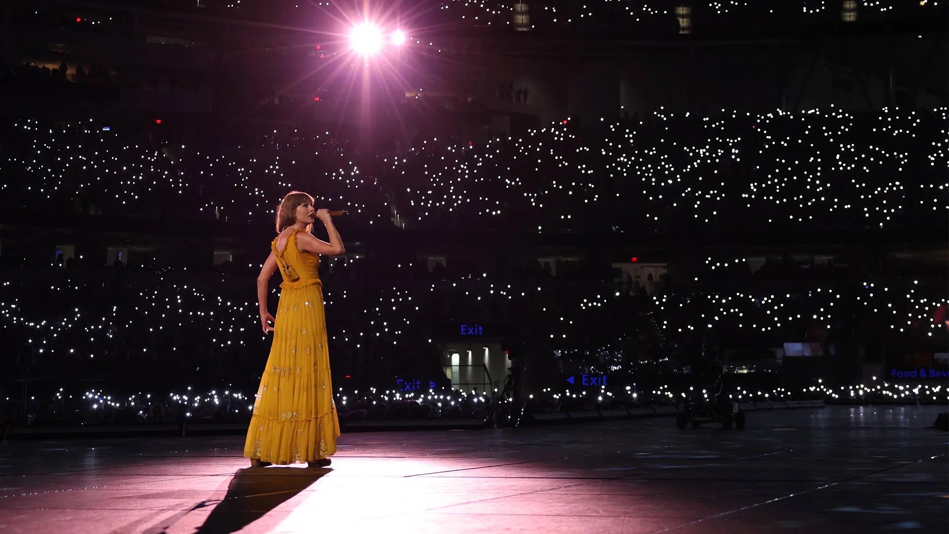 Taylor Swift wearing yellow dress singing into microphone infront of a crowd of thousands of fans holding bright lights