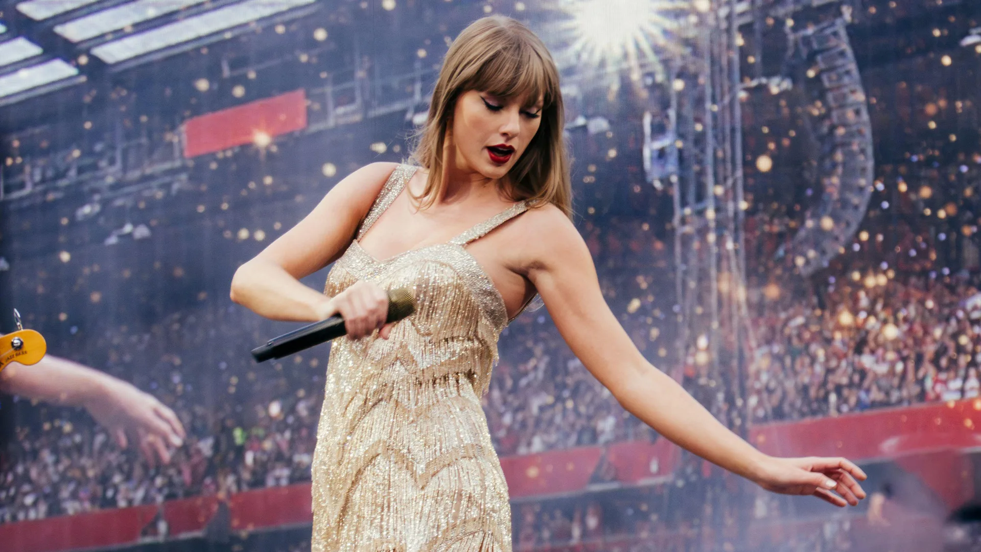 A photograph of Taylor Swift at her Eras concert tour wearing a gold beaded fringe dress holding a mic with her arm stretched out looking down against a stadium crowd background