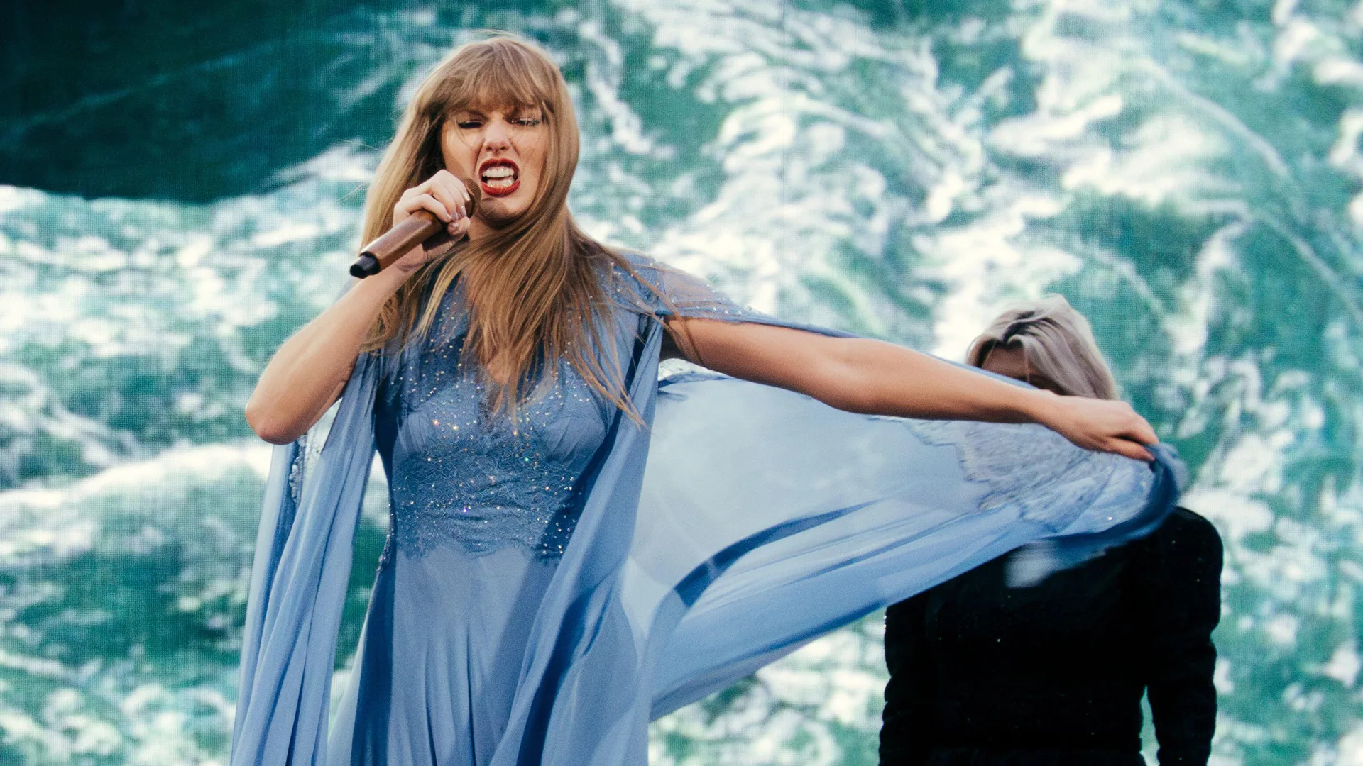 A photograph of Taylor Swift wearing a blue dress with long flowing sleeves signing with her teeth bared into a mic against a blue background. A blonde woman stands behind her.