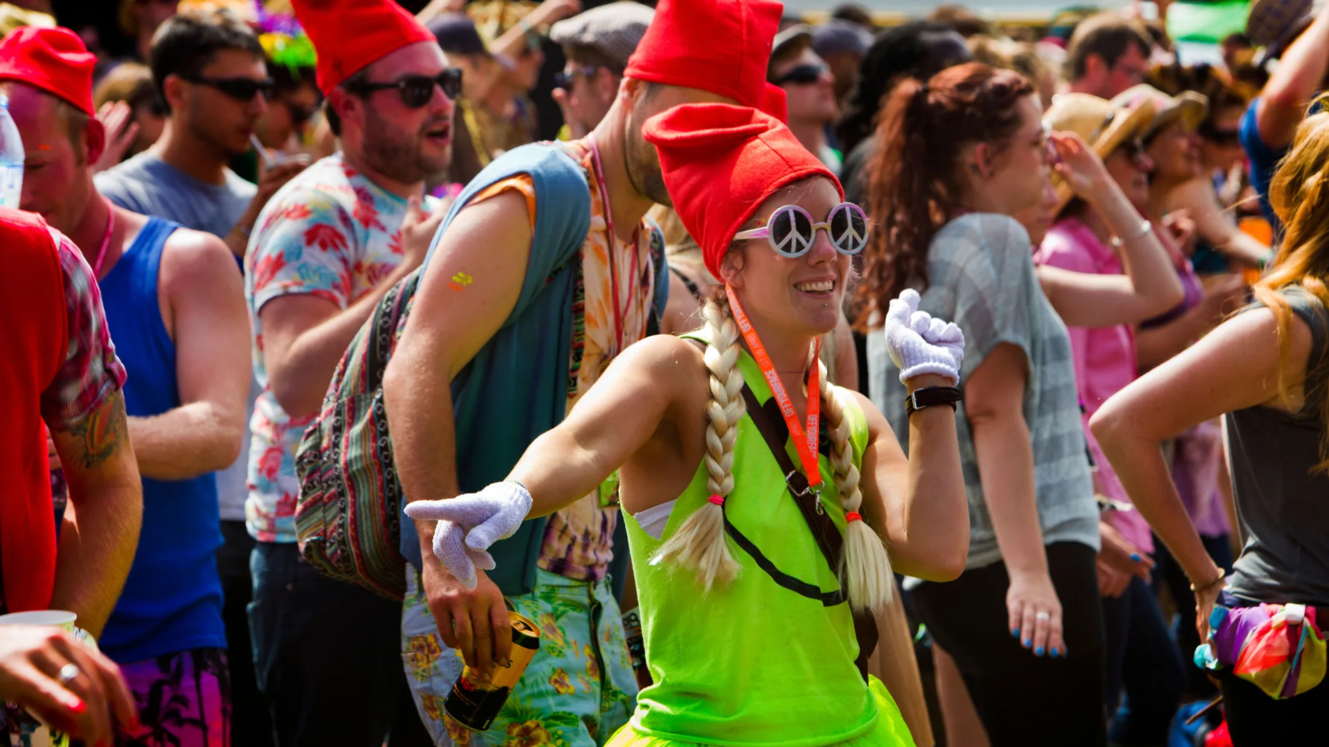 A photo of a woman wearing a green dress, peace sign sunglasses and a red hat dancing with white gloved hands pointing - around her is a festival crowd