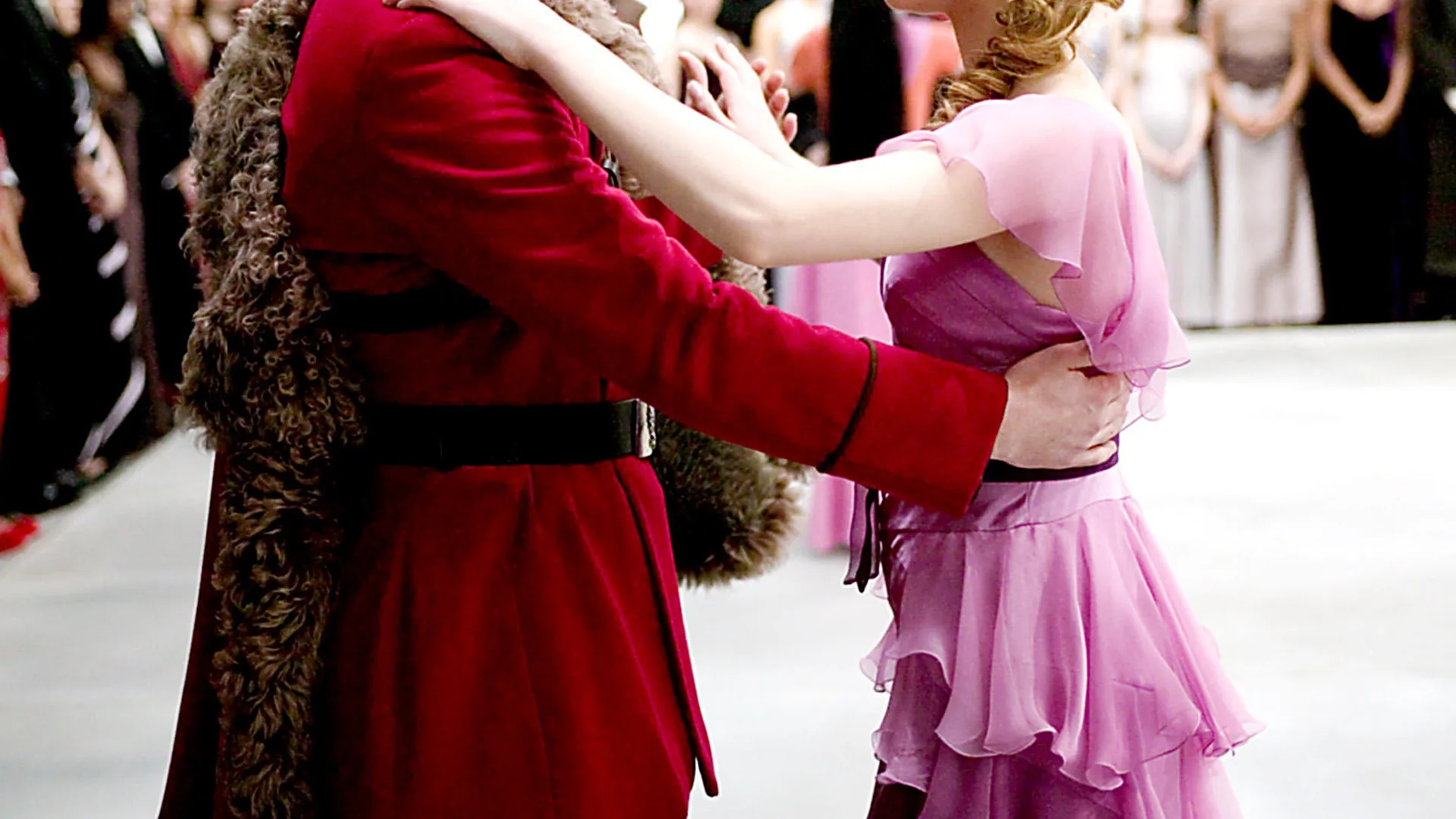 A photograph of a couple dancing. The man is wearing a red velvet coat and the girl is wearing a pink floaty dress.