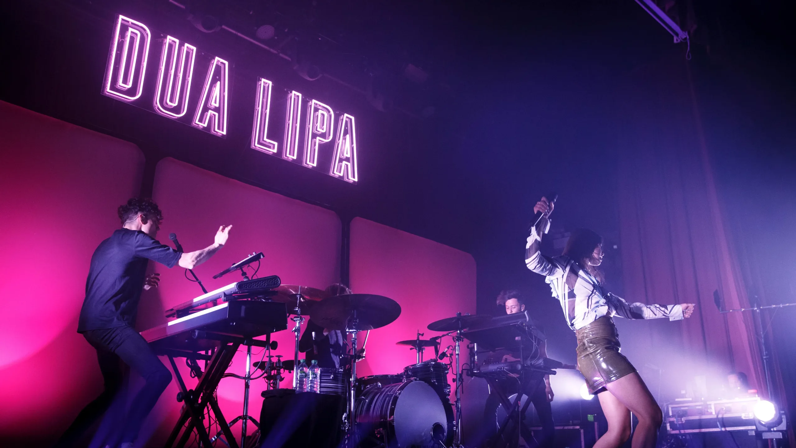 Dua Lipa performs with band on stage against a pink backdrop