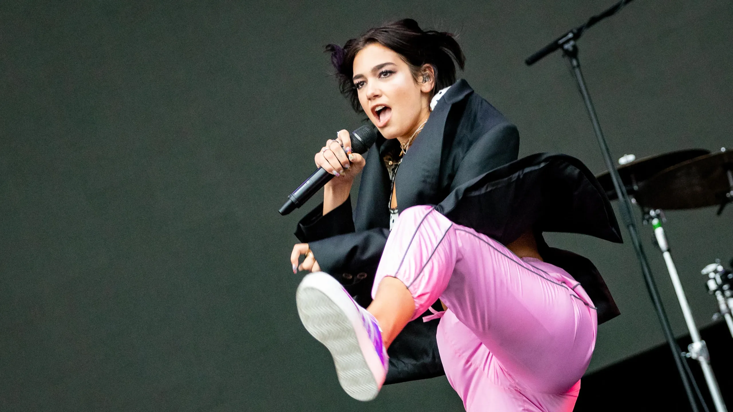 Dua Lipa performs highkick on stage wearing black jacket and pink trousers