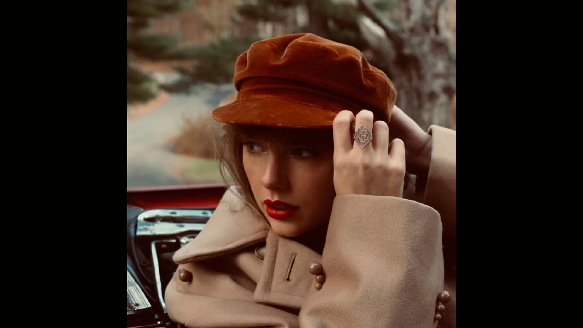 Taylor Swift's album RED - Taylor's Version showing the singer wearing a red hat and beige coat looking to the side holding the peak of her hat in her hand with a car and road out of focus behind her.