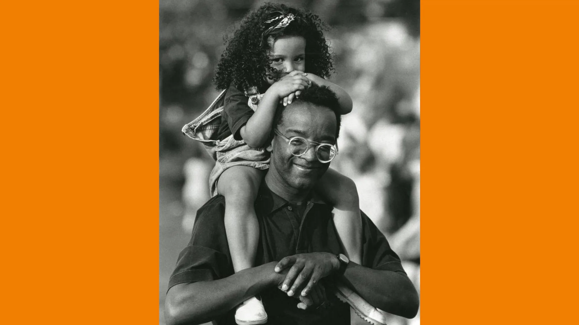 A photograph of a man wearing glasses and a black top holding his daughter on his shouders. She has her head down on her hands which are on top of his head and both are smiling.