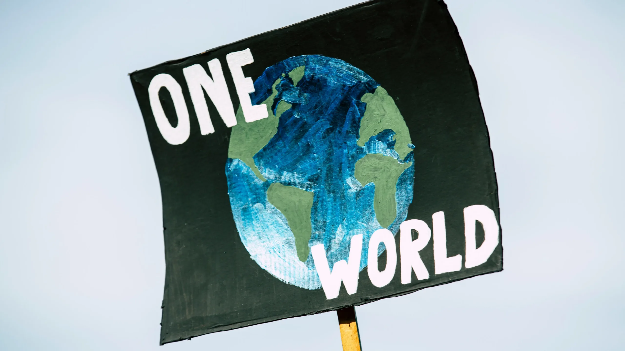 A sign held up against the sky with an illustration of planet earth against a black background, accompanied by the words 'ONE WORLD' in white capital letters