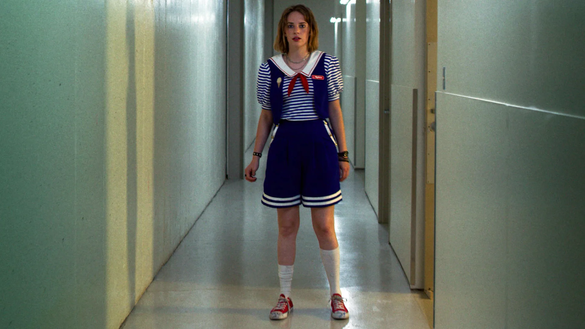 A photograph of the character Robin from Stranger Things stood in a corridor wearing a sailor outfit looking nervous