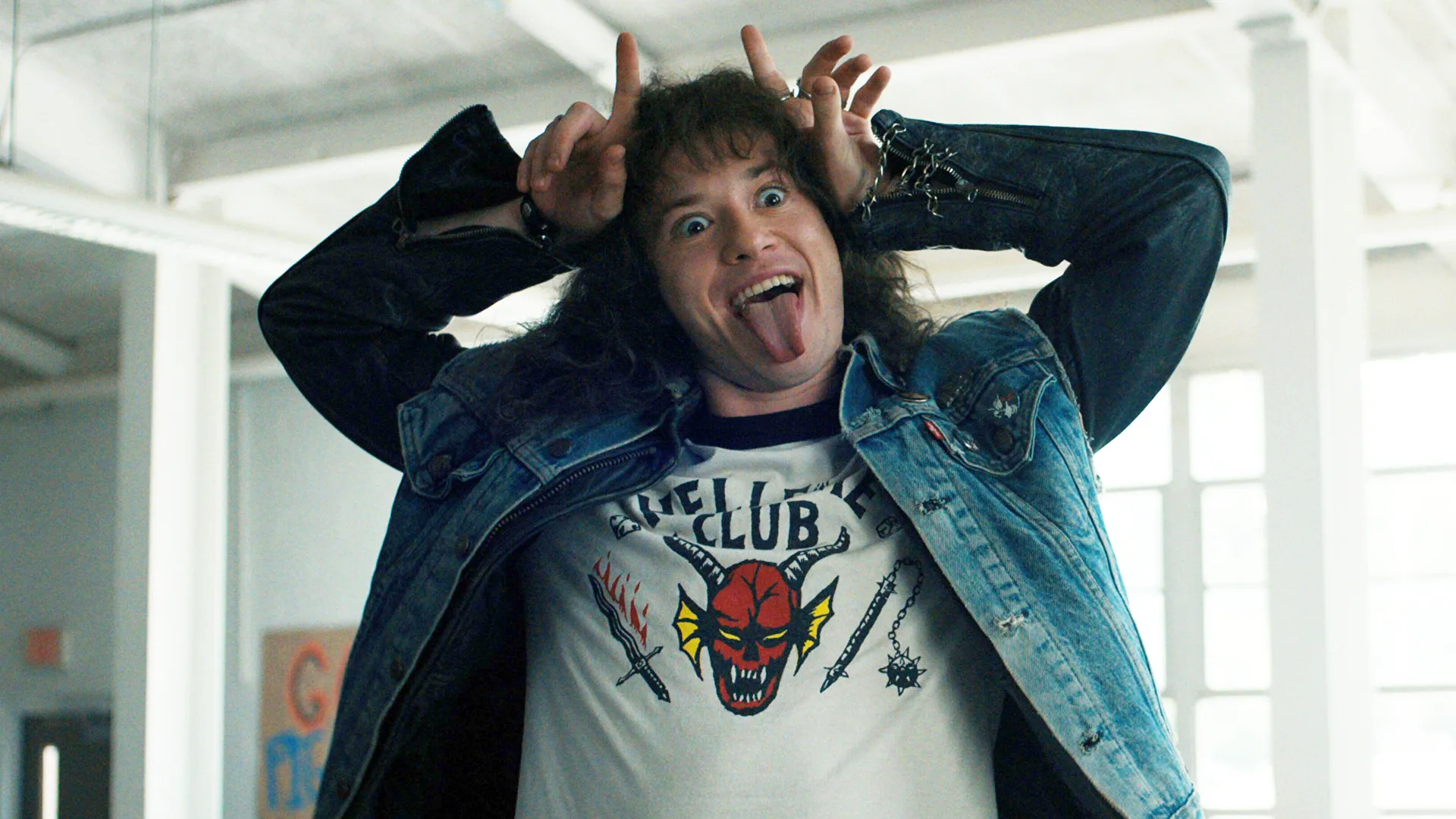 A photo of Eddie Munson in Stranger Things wearing a Hellfire tshirt holding his hands up to his head and making a devil sign with his fingers whilst sticking his tongue out