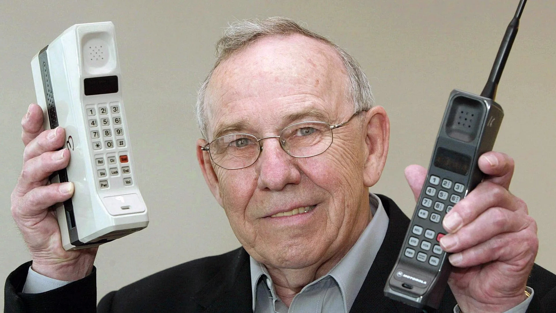 A photograph of Rudy Krolopp the Motorola design chief who made the first mobile phone - he is holding both original models up with a smile on his face against a beige background
