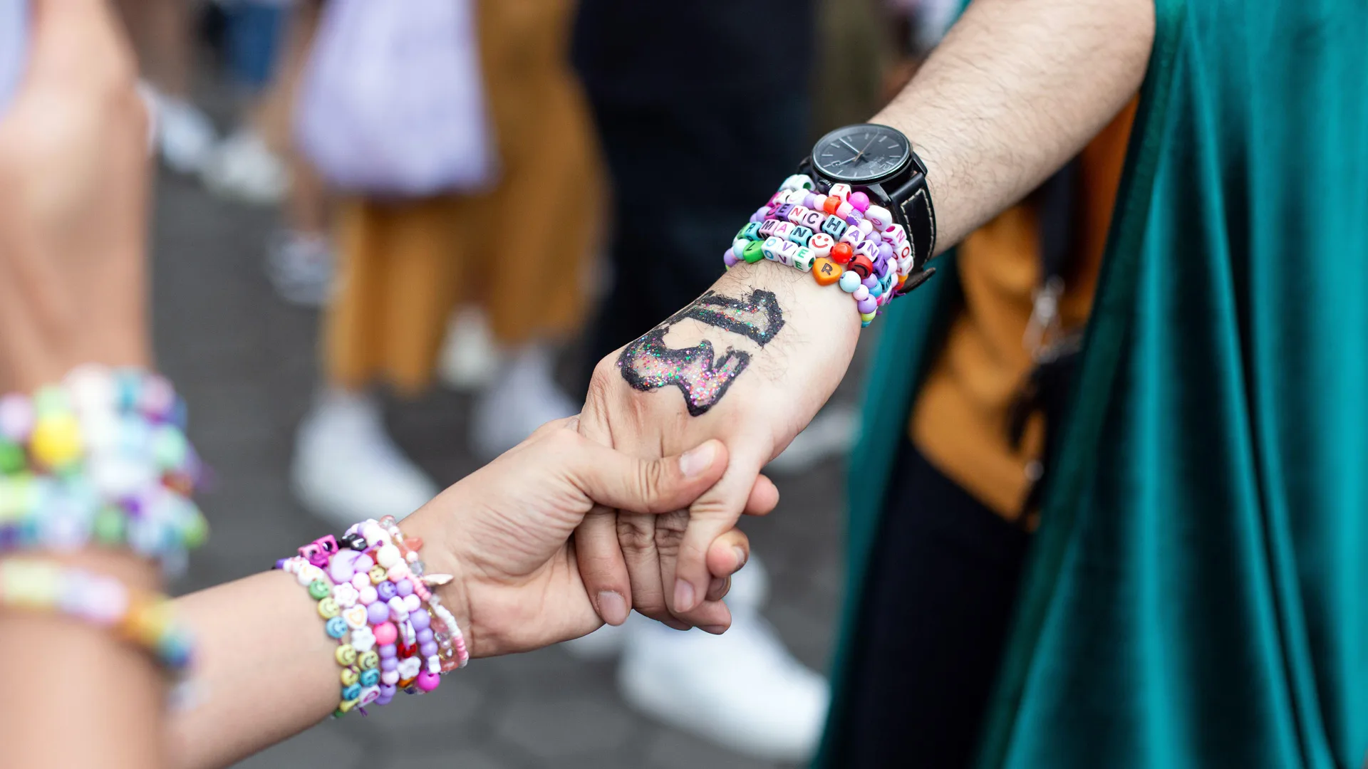 A photograph of two hands holding with Taylor Swift beaded bracelets on each arm and the hand facing up has a painted number 13 in black and glitter