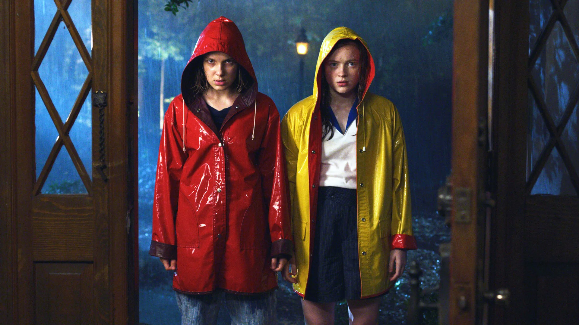 A photo of Max and Eleven from Stranger Things stood in the rain wearing shiny plastic raincoats of yellow and red